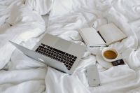 Macbook, an iPhone, a coffee, a chocolate and a notebook in a bed