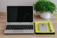 Silver Acer laptop, a white Apple iPhone, a yellow notebook and a green plant on a wooden desk