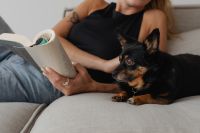 A woman reads a book on the couch with her dog